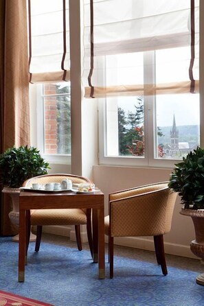 A room with a dining table set for two, two chairs, a window with curtains, and a view of a building with a spire.