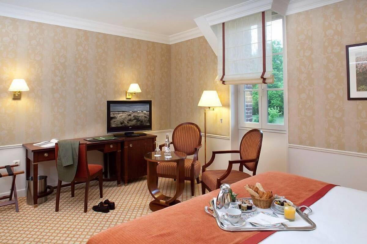 A hotel room with a bed, a desk, two chairs, a television, and a window with a view of greenery.