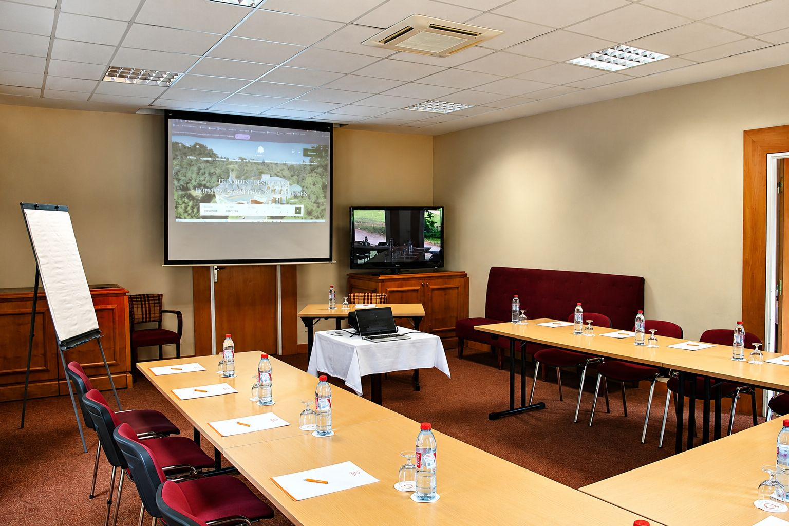 Une salle de conférence équipée d’un écran de projection, d’un téléviseur et d’un tableau blanc. On y trouve des tables avec des chaises et des bouteilles d’eau.