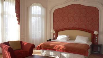 A hotel room with a large bed, a red armchair, a small table, and a window with sheer curtains.