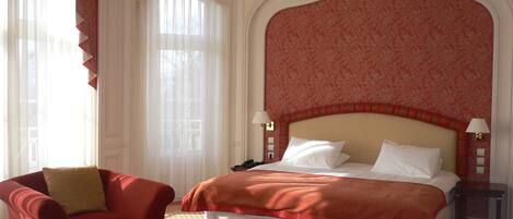 A hotel room with a large bed, a red armchair, a small table, and a window with sheer curtains.
