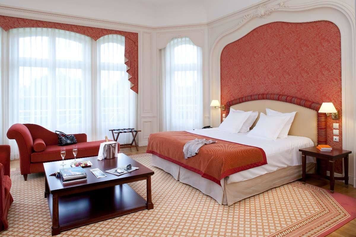 A hotel room with a large bed, a red sofa, a wooden coffee table, and a small side table with a lamp.