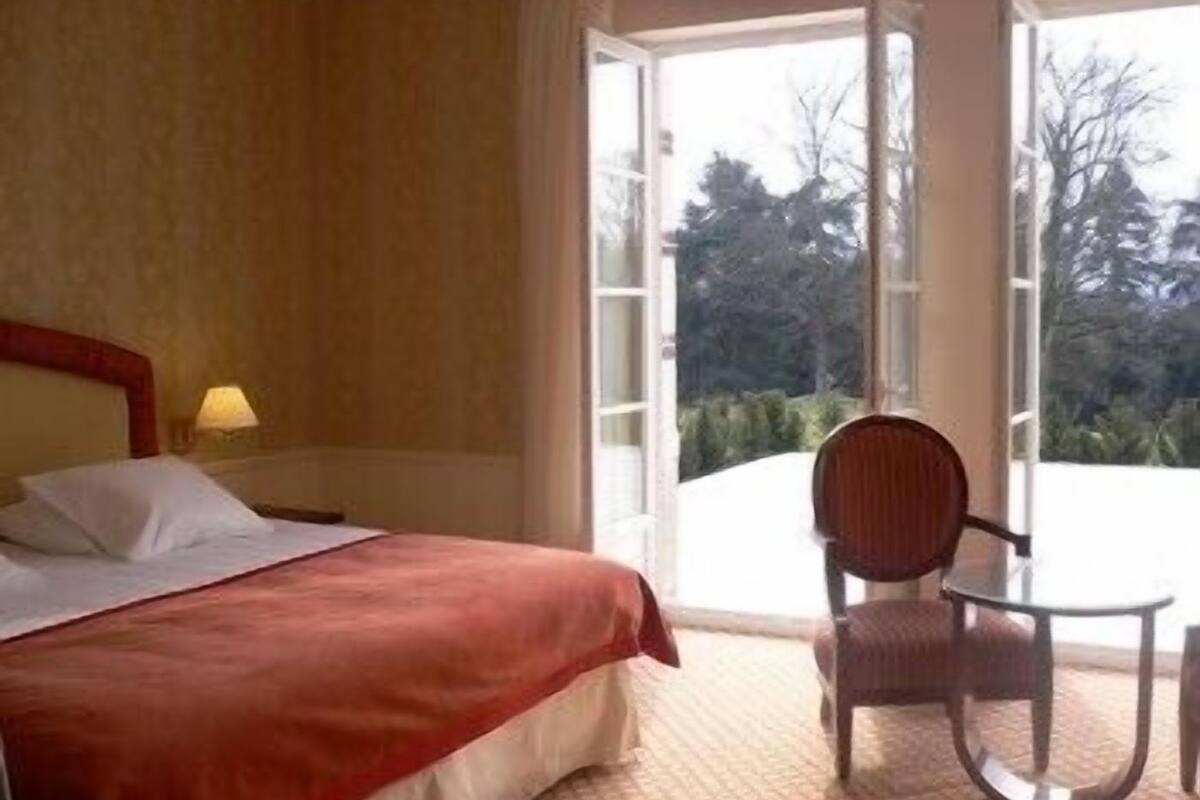 A hotel room with a bed, a chair, and a table near a window with a view of trees.
