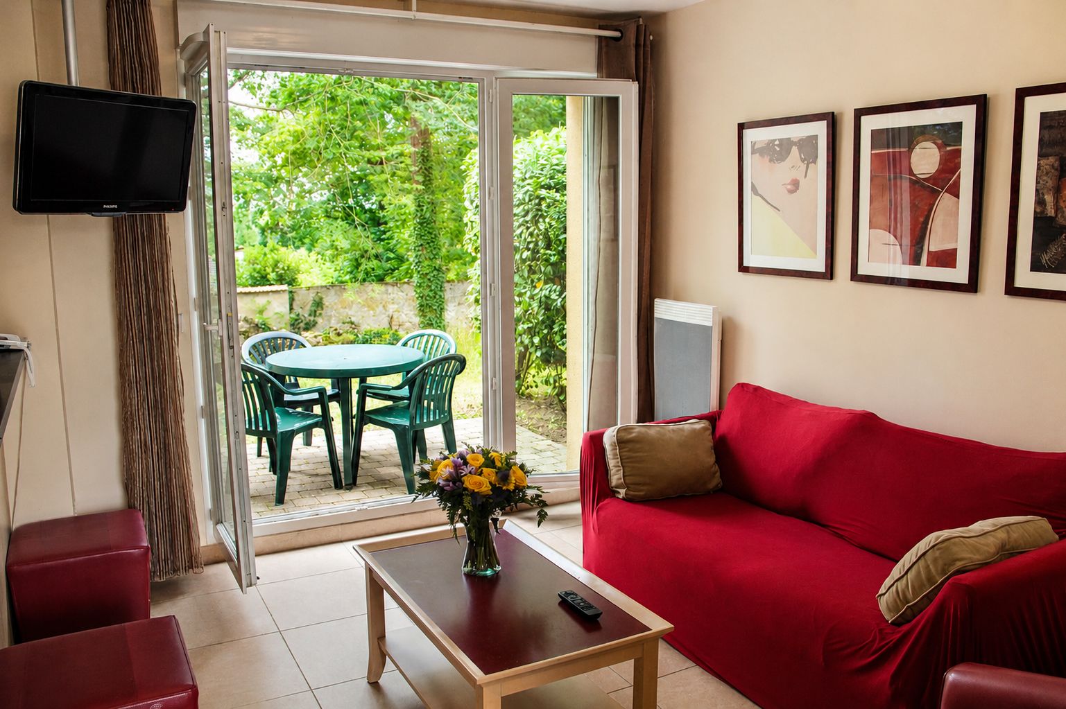 A living room with a red sofa, a coffee table with a vase of flowers, a television mounted on the wall, and a sliding glass door leading to an outdoor area with a table and chairs.