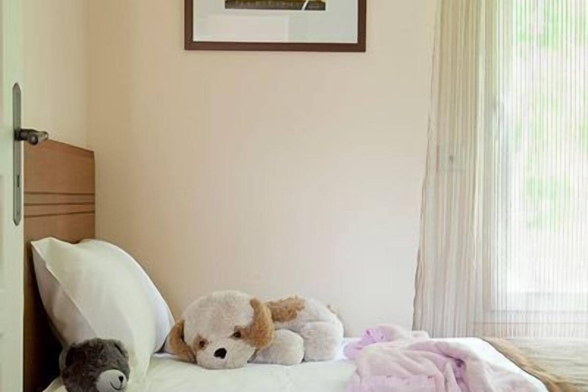 A bedroom with a bed, pillows, a teddy bear, a framed picture, and a window with curtains.