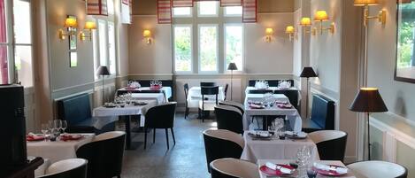 A restaurant with multiple tables set for dining, featuring a classic interior design with wooden beams on the ceiling and natural light from large windows.