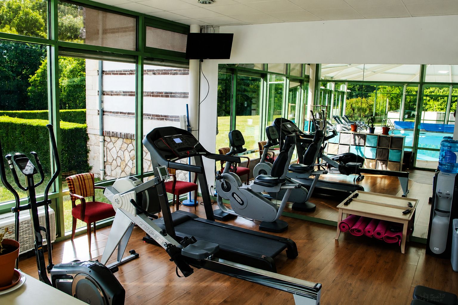 Un gimnasio con máquinas de cardio, una máquina de remo y una banca. Hay un ventanal con vistas a zonas verdes y una piscina.