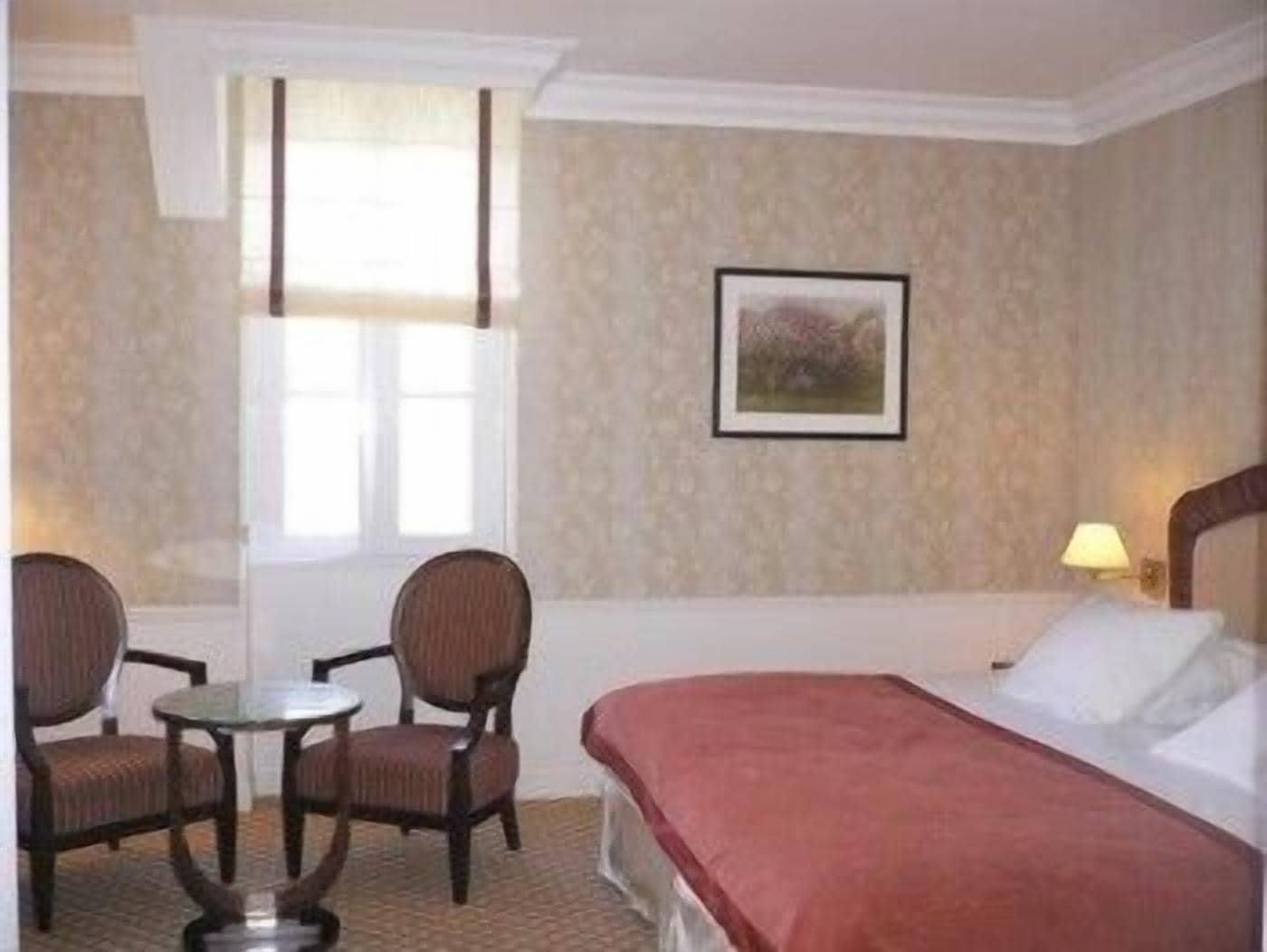 Habitación de hotel con cama, dos sillas, una mesita, una ventana y una lámpara de pared.