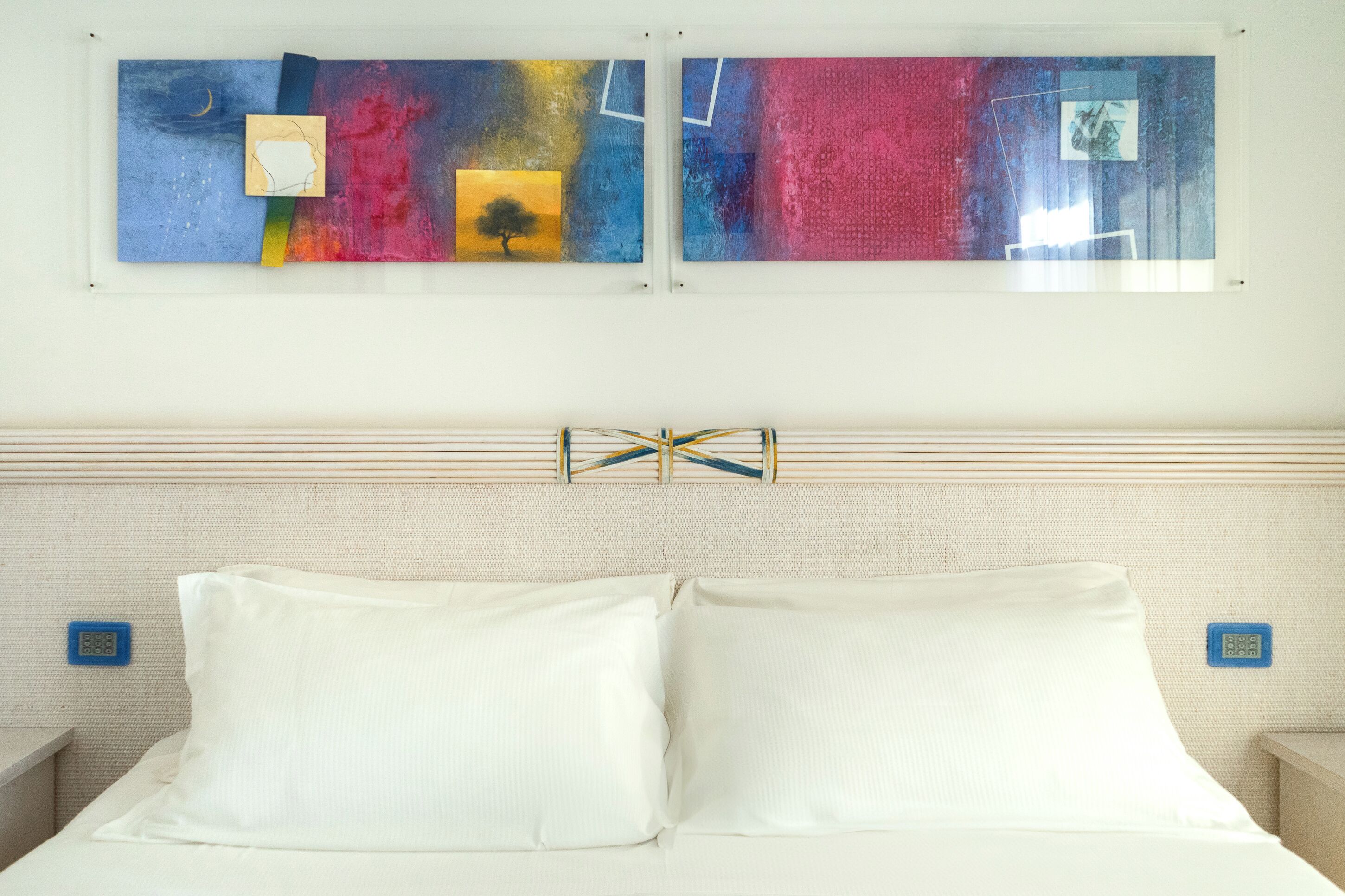 A hotel room with a bed, white pillows, a headboard with a geometric design, and abstract artwork on the wall.