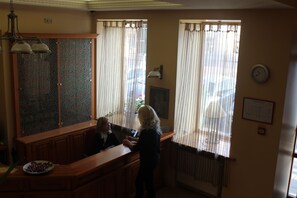 A reception area with a wooden counter, a clock on the wall, and a window with curtains. - Alexa Old Town (Vilnius)