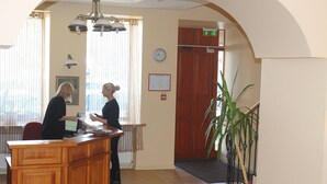 A reception desk with two staff members attending to guests, a clock on the wall, and a staircase leading to an upper floor. - Alexa Old Town (Vilnius)