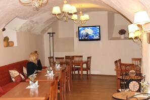 A dining area with a long wooden table, multiple chairs, a red sofa, and a television mounted on the wall. - Alexa Old Town (Vilnius)