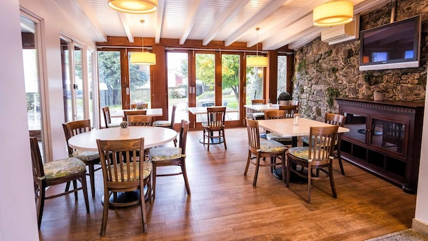 Un espace repas avec des tables et des chaises en bois, de grandes fenêtres et un mur en pierre agrémenté d’une cheminée.
