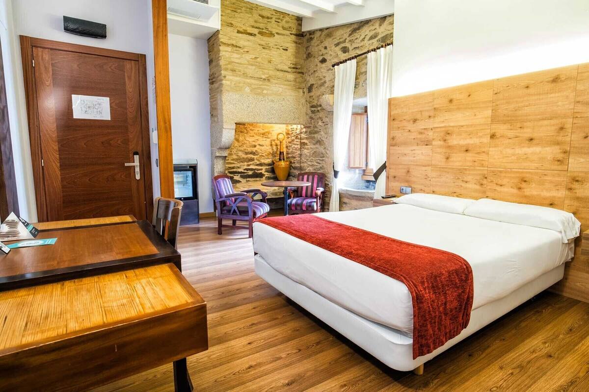 A hotel room with a wooden headboard, a bed with a red blanket, a wooden desk, a chair, and a stone wall.