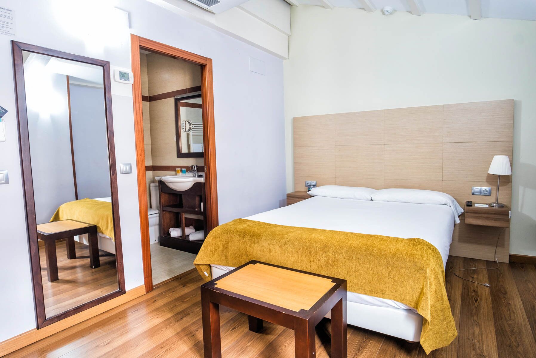 Habitación de hotel con cama, mesita de noche de madera, lámpara de noche, una pequeña mesa, un espejo y un baño visible a través de una puerta abierta.