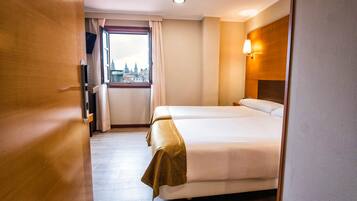 A hotel room with two beds, a window with a city view, a wooden headboard, and a flat-screen TV.