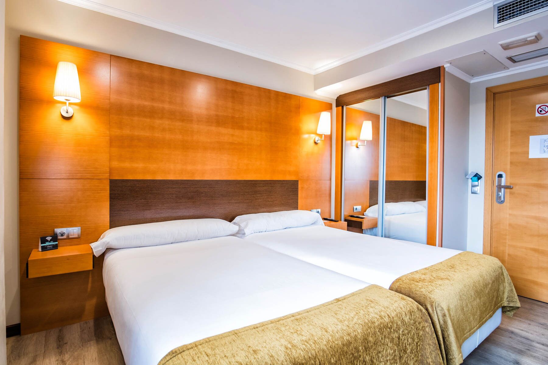 Habitación de hotel con una cama grande, cabecera de madera, lámparas de noche, un espejo y una puerta con un cartel de 'No Fumar'.
