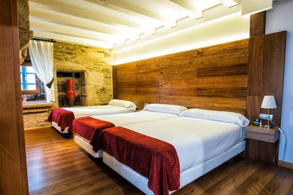 A hotel room with two beds, wooden flooring, a stone wall, and a modern ceiling light fixture.