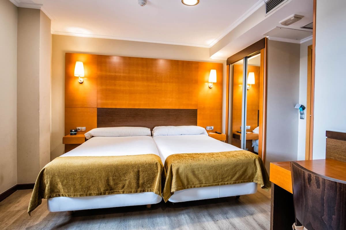 A hotel room with two beds, wooden headboards, bedside lamps, a large mirror, and a wooden dresser.