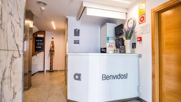Una recepción con un cartel que dice "Benvidos!" en un vestíbulo moderno con suelo de mármol, una estatua y un reloj en la pared.