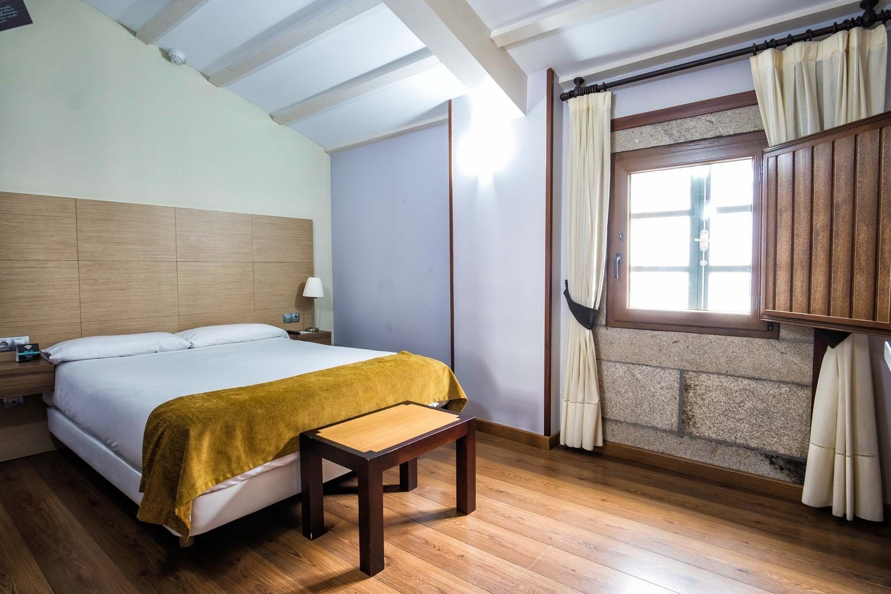 A hotel room with a wooden bed, a bedside table, a nightstand, a window with curtains, and a view of the outside.