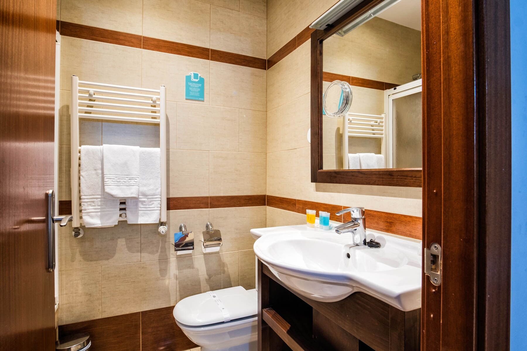 A bathroom with a white sink, a mirror, and a toilet. There are towels hanging on a rack and a sign on the wall.
