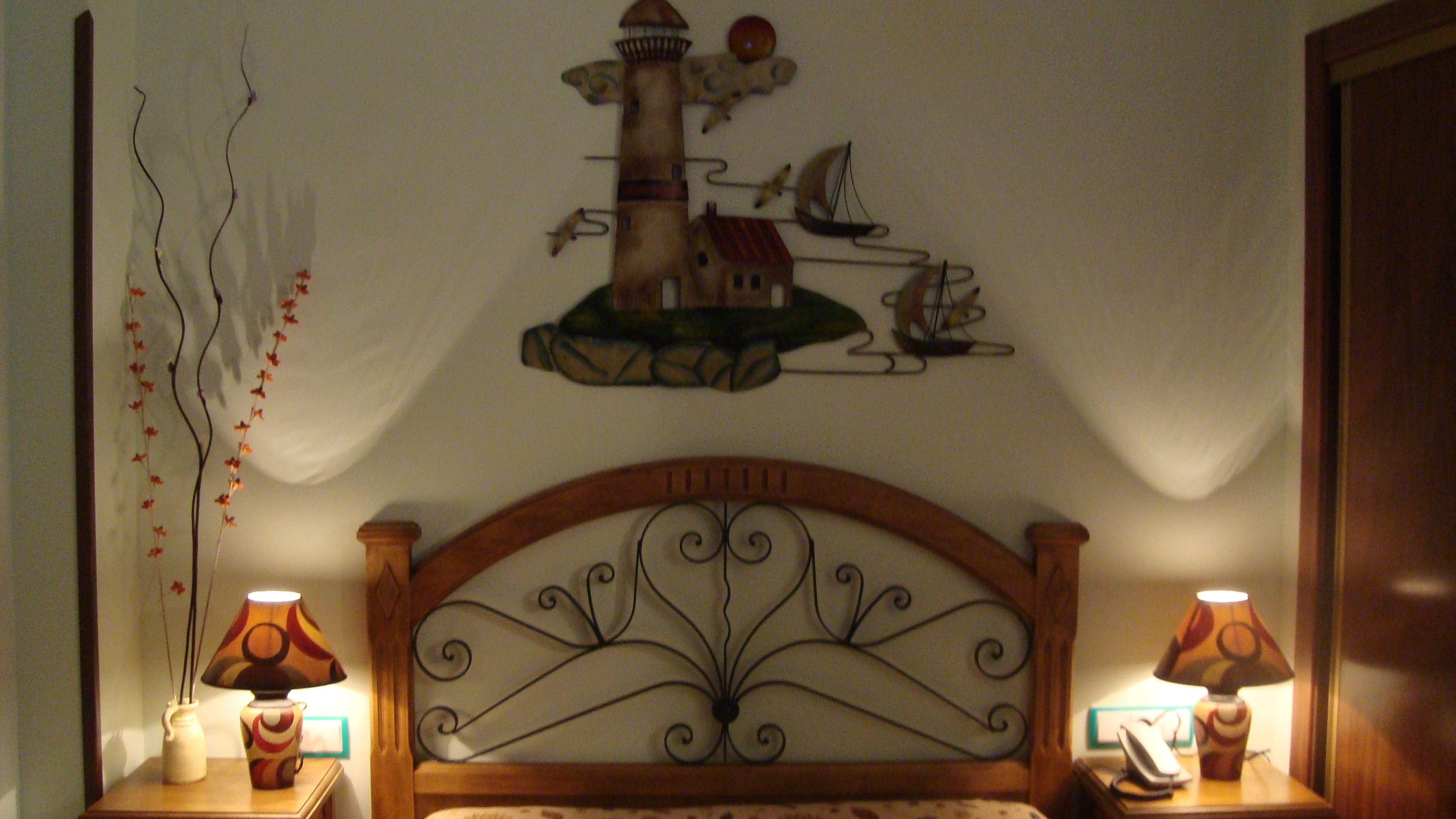 A bedroom with a decorative headboard, two bedside lamps, a wall-mounted artwork of a lighthouse, and a vase with branches on the nightstand.