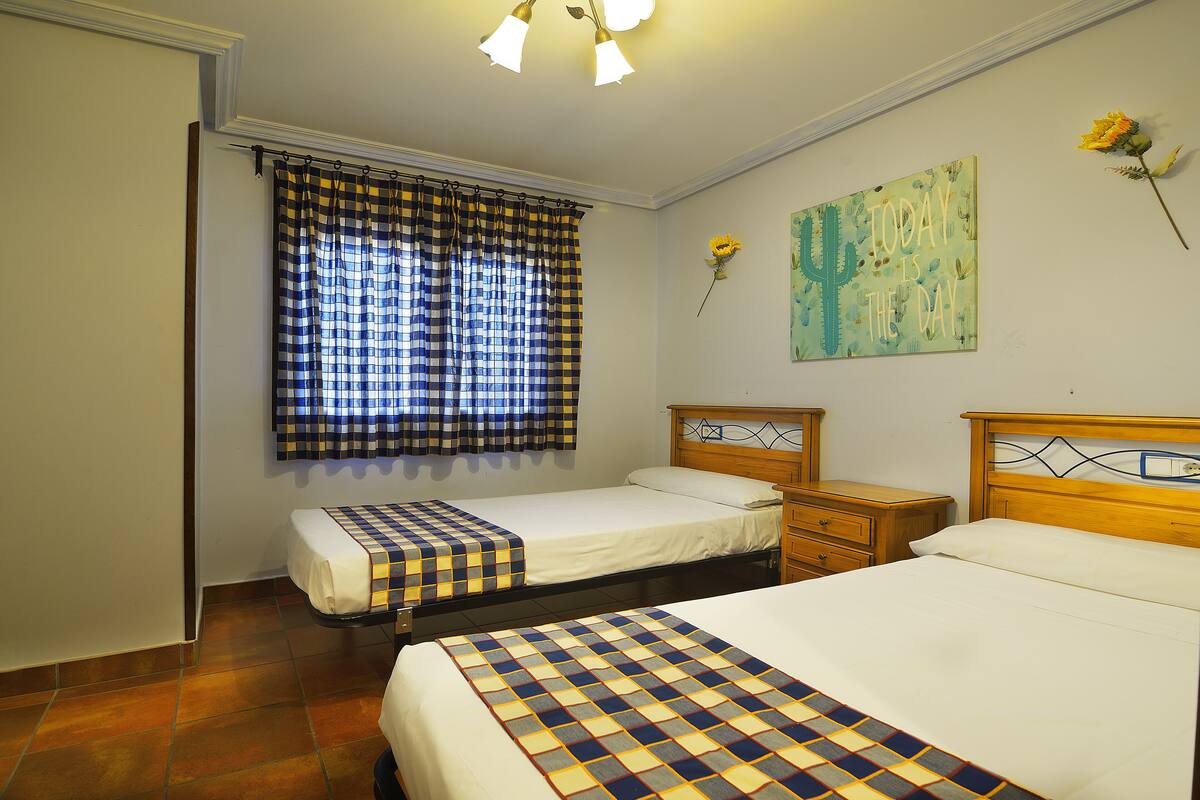 Quarto de hotel com duas camas, uma janela com cortinas xadrez, mesa de cabeceira, cabeceira de madeira e um objeto decorativo na parede.