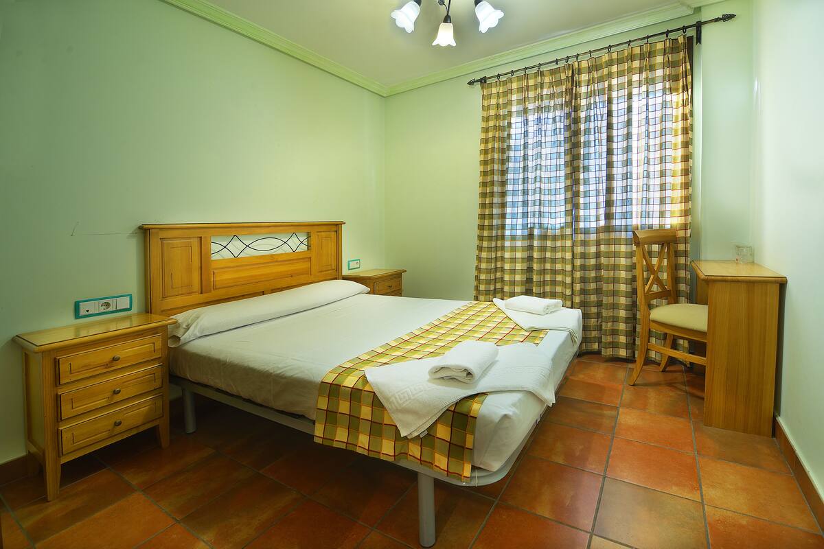 Uma cama bem arrumada com colcha xadrez, uma mesa de cabeceira de madeira com gavetas, uma cadeira e uma pequena mesa com abajur.