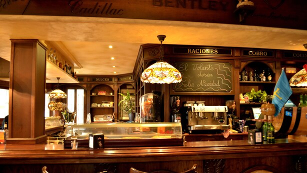 A bar area with a wooden counter, bar stools, a chalkboard menu, and a decorative stained glass light fixture. - El Volante (Ciempozuelos)