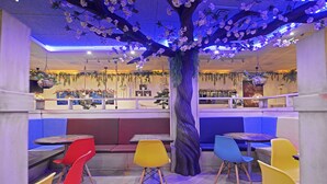 A restaurant interior with a central tree decoration, colorful chairs, and a counter area. - El Volante (Ciempozuelos)