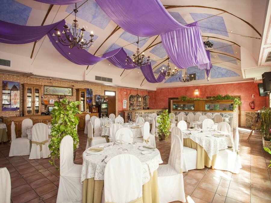 Un salón de banquetes con mesas redondas dispuestas para comer, sillas blancas y cortinas moradas.
