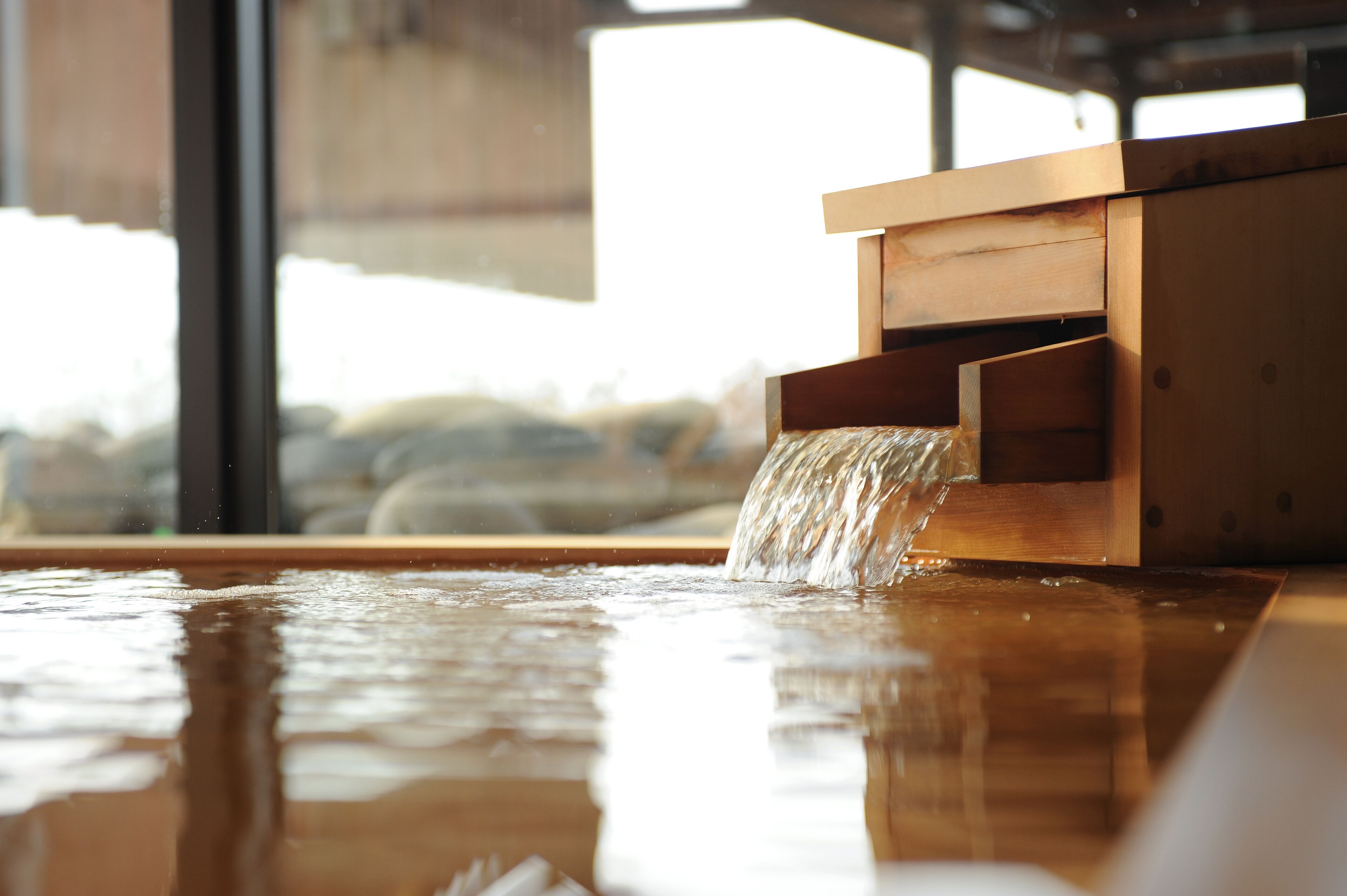 A wooden water feature with a stream of water flowing into a basin, set against a backdrop of large windows.