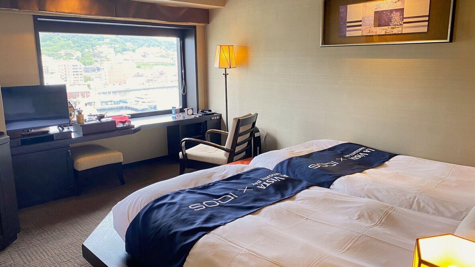 A hotel room with a bed, a desk, a chair, a TV, and a window with a view of buildings.