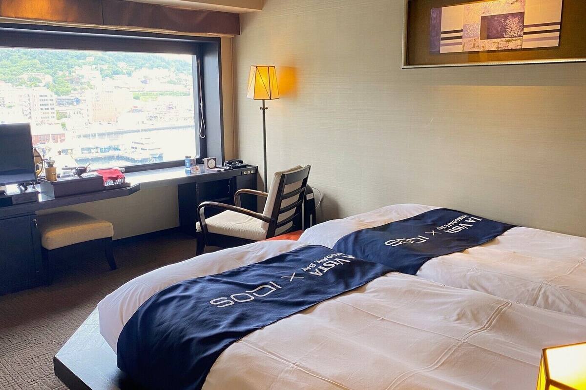 Habitación de hotel con cama, escritorio, silla, televisor y ventana con vistas a edificios.