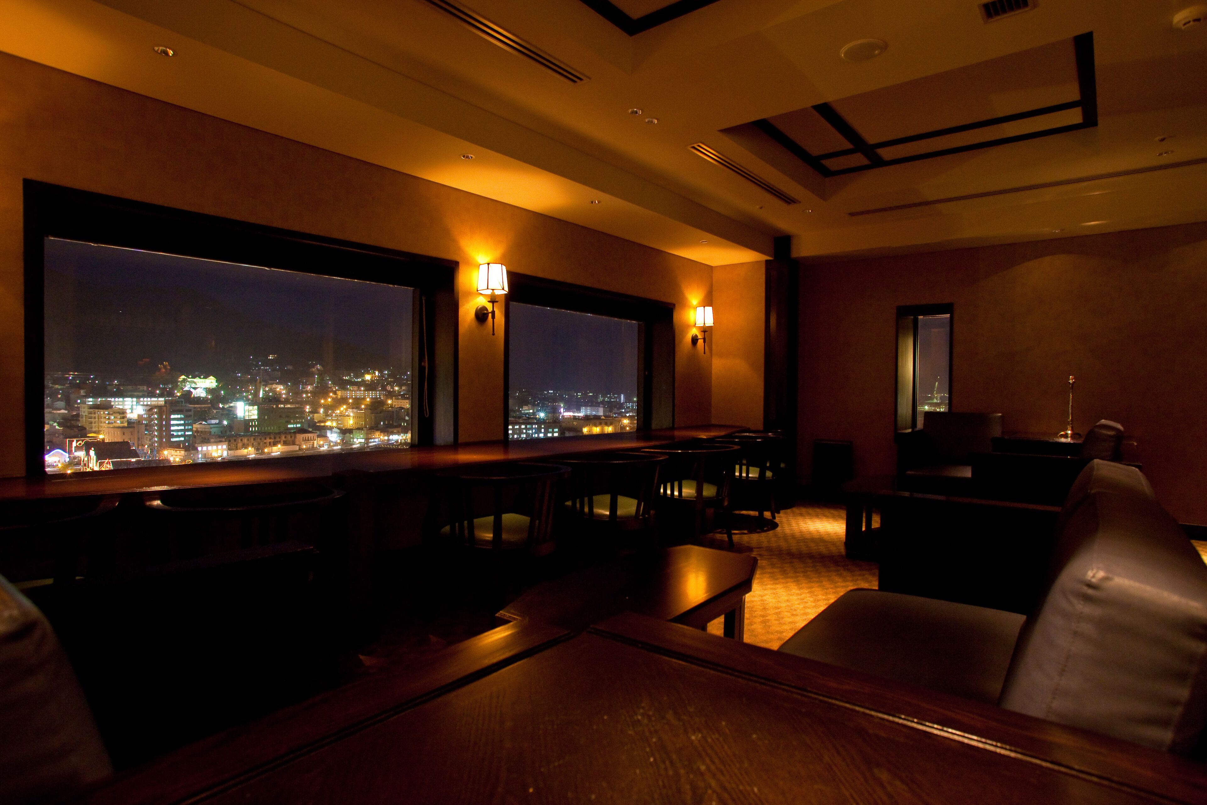 A dimly lit room with a large window overlooking a cityscape at night. There are several tables and chairs arranged for dining or socializing.