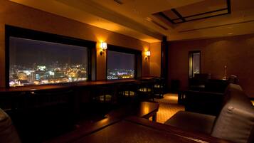 A dimly lit room with a large window overlooking a cityscape at night. There are several tables and chairs arranged for dining or socializing.