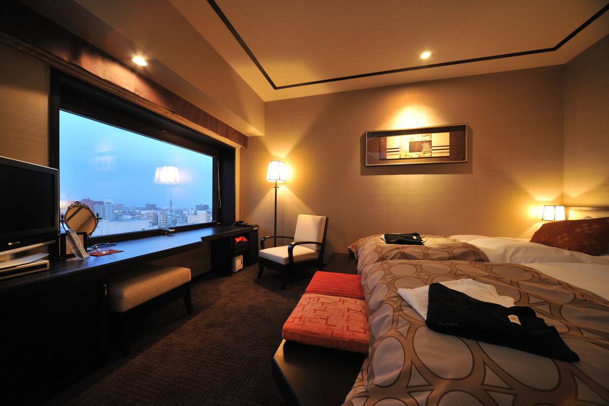 Habitación de hotel con cama, escritorio con televisor, una silla y ventana con vista a la ciudad.
