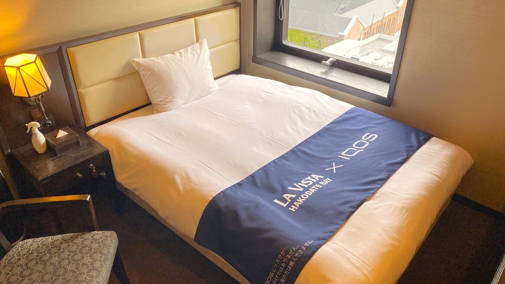 A hotel room with a bed, a nightstand, a lamp, and a window with a view.