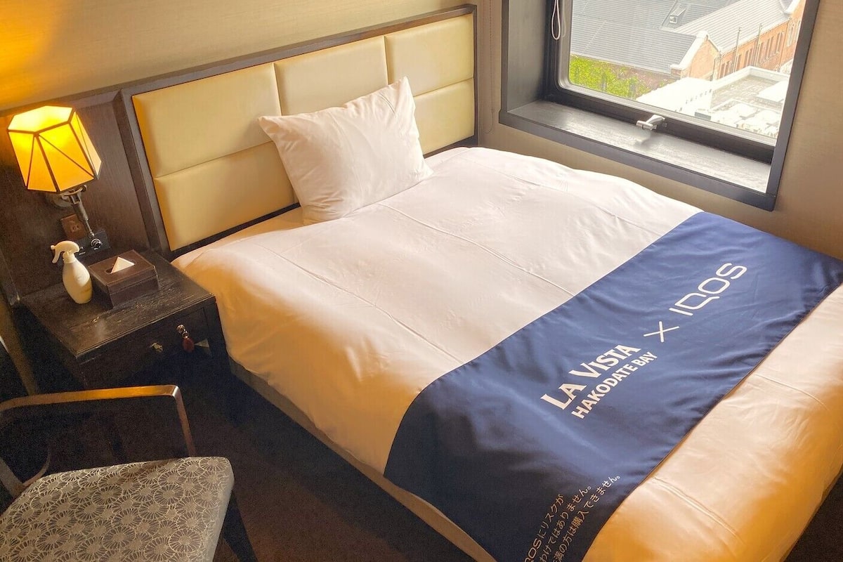 Una habitación de hotel con cama, mesita de noche, lámpara y ventana con vistas.