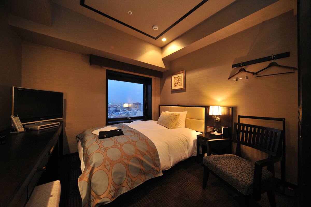 Habitación de hotel con cama, escritorio con televisor, silla, ventana con vista a la ciudad y perchero en la pared.