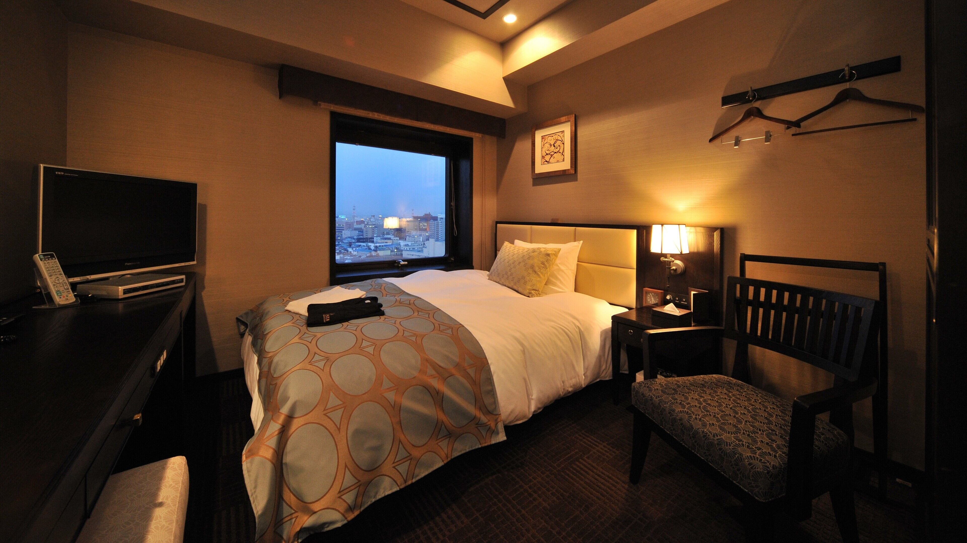 A hotel room with a bed, a desk with a TV, a chair, a nightstand, and a window with a city view.