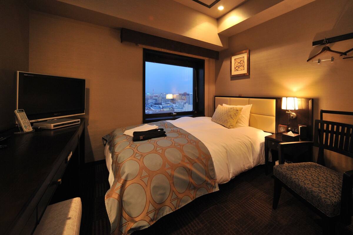 Habitación de hotel con cama, escritorio con televisor, silla, mesita de noche y ventana con vista a la ciudad.