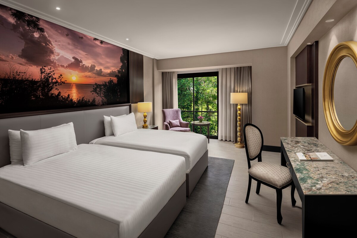 A hotel room with two beds, a desk, a chair, a TV, and a large window with a view of greenery.
