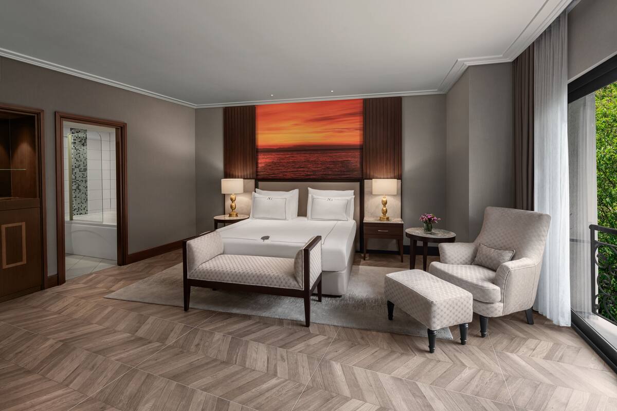 Ein Hotelzimmer mit einem großen Bett, zwei Nachttischen mit Lampen, einem Sessel, einer Couch und Blick ins Freie.