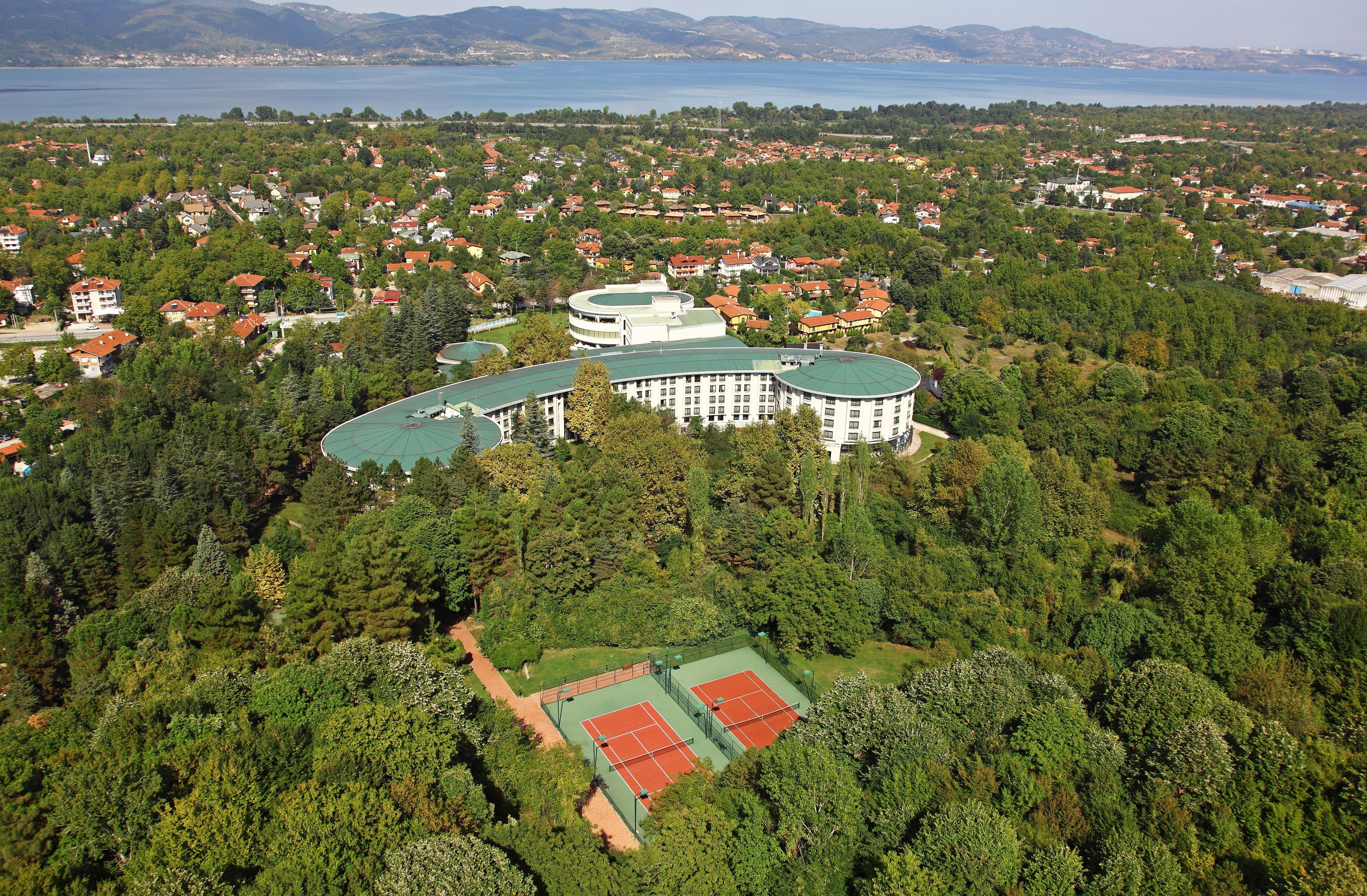 A large, multi-story building surrounded by dense greenery and a tennis court.
