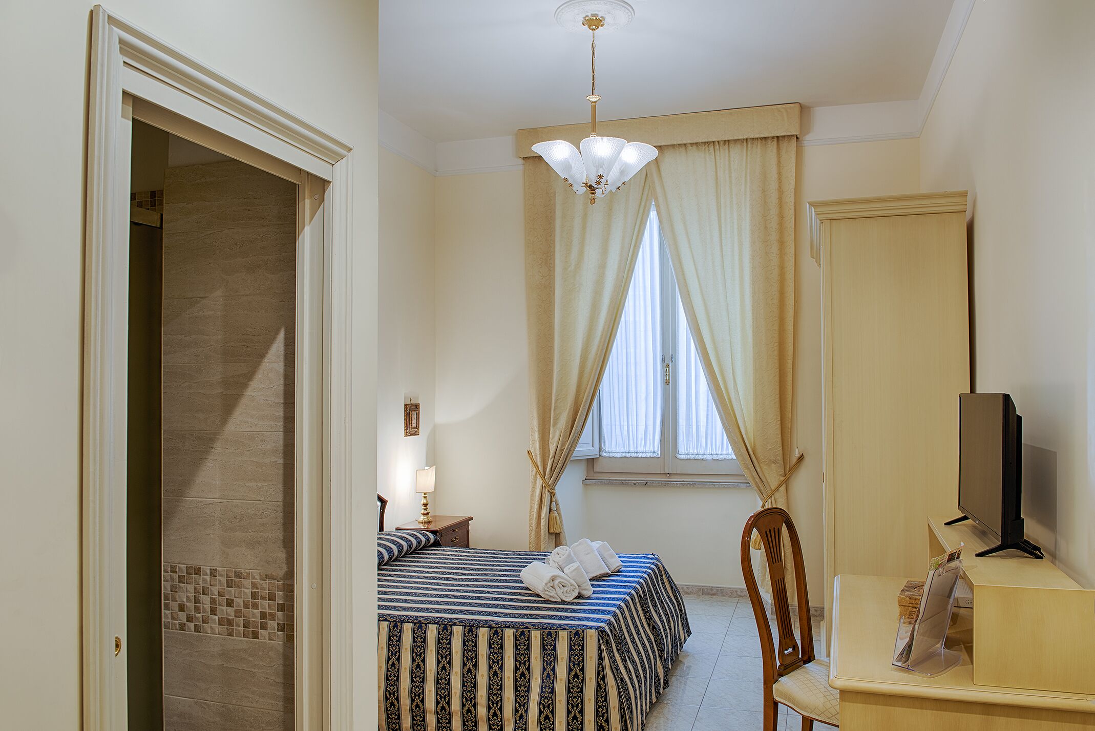 Habitación de hotel con cama, escritorio con televisor, silla y ventana con cortinas.