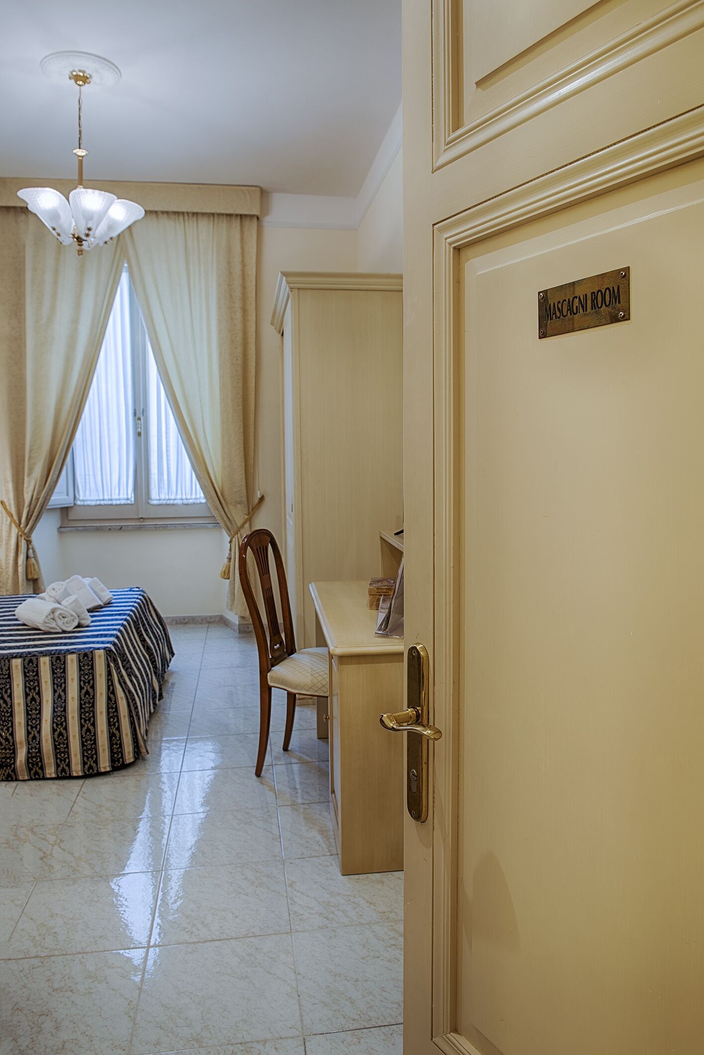 Una habitación de hotel con cama, escritorio, silla, candelabro y una puerta con la leyenda 'MASCAGNI ROOM'.