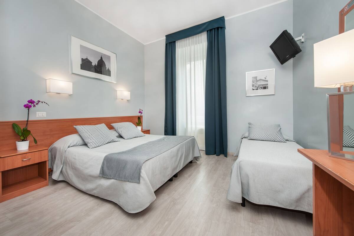 A hotel room with two beds, a nightstand, a bedside table, a potted plant, and a wall-mounted TV.