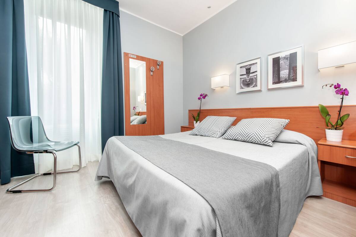 A hotel room with a bed, a chair, a nightstand, a mirror, and two wall-mounted lamps.